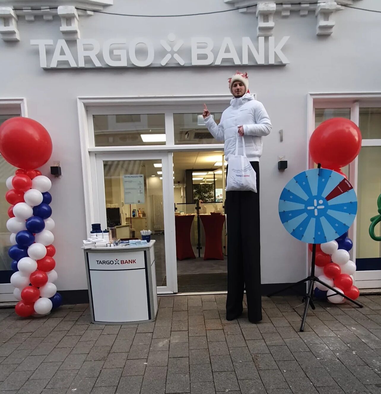 Stelzenläufer in weißer Jacke mit Give-away-Tasche vor der neu eröffneten Targobank-Filiale in Heide, flankiert von Ballonsäulen und Glücksrad